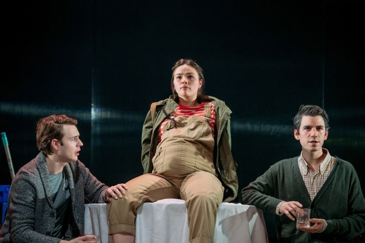 Actor made to look pregnant sits on a table